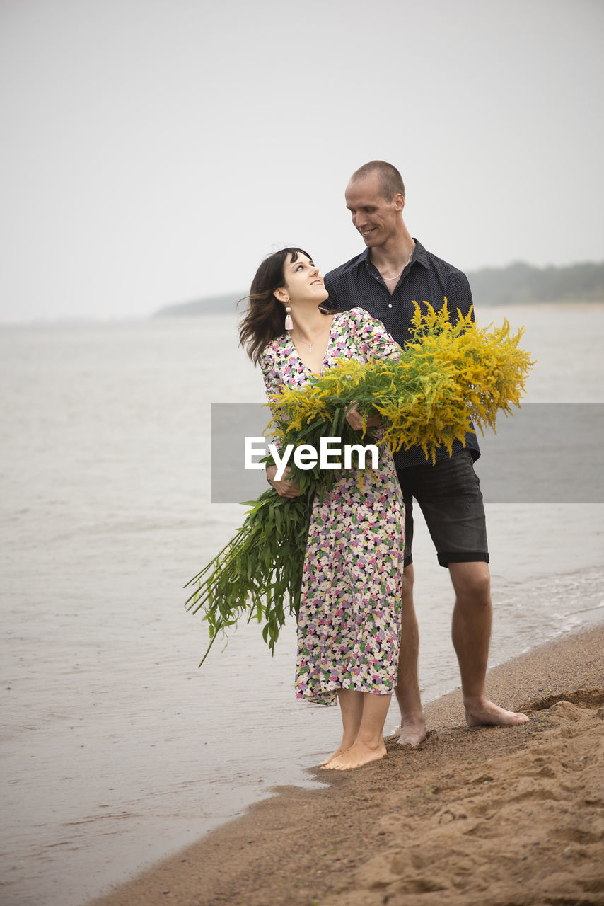 Young romantic couple with big bouquets of wild flowers posing on the beach