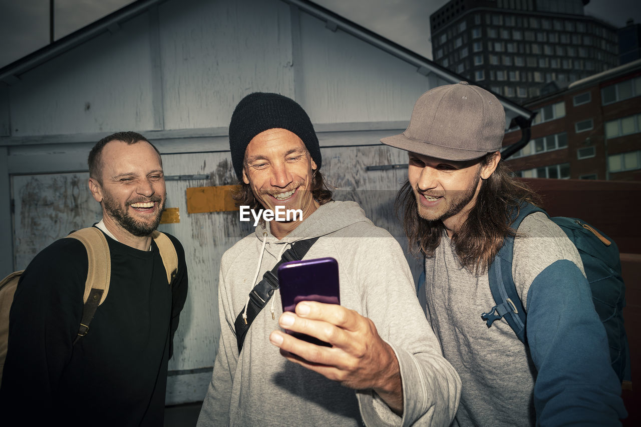 Happy male friends taking selfie on smart phone against shed on street