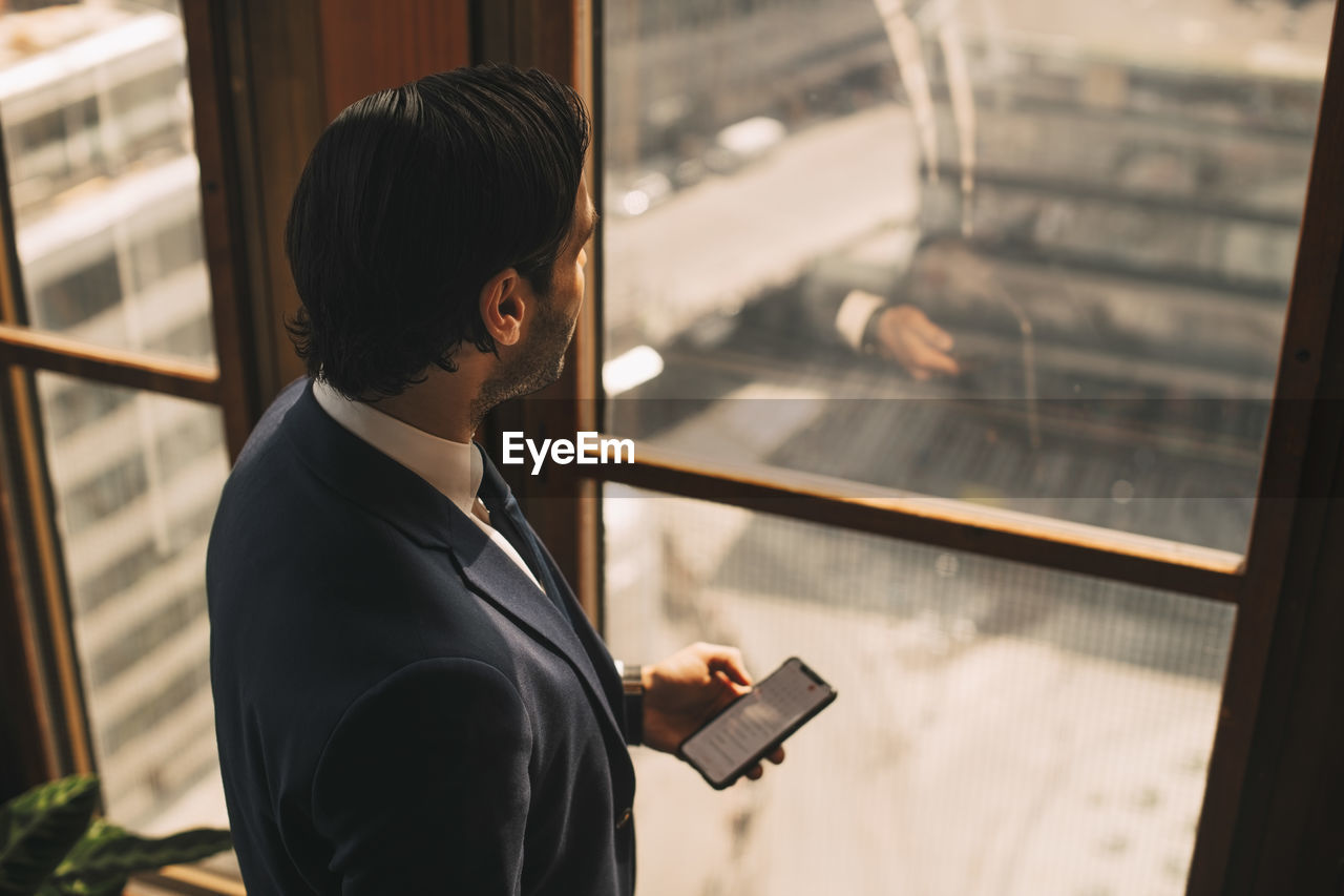 High angle view of thoughtful financial advisor texting on mobile phone by window at office