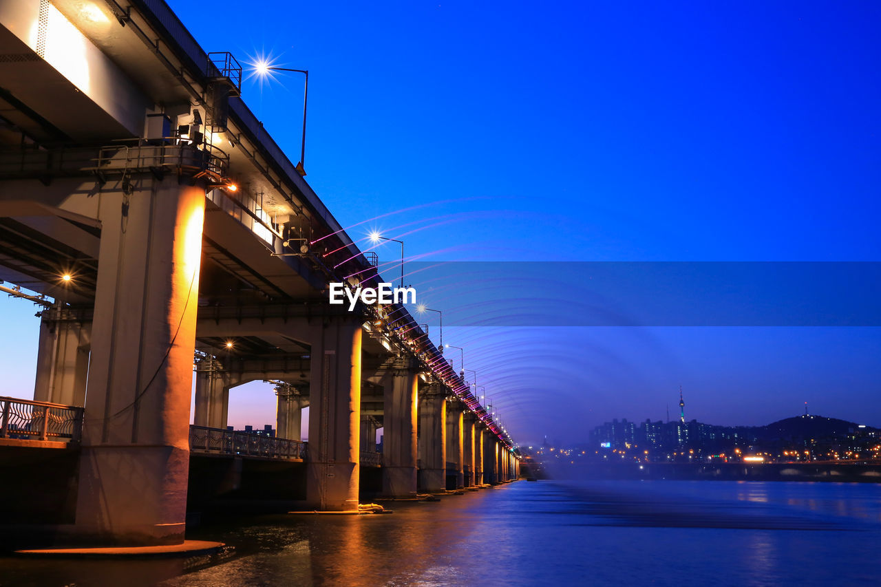 Illuminated bridge over river against blue sky at night