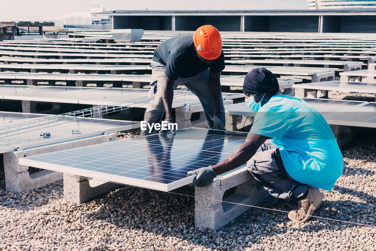 Busy black male engineers installing solar batteries in construction site of modern factory during sunny day
