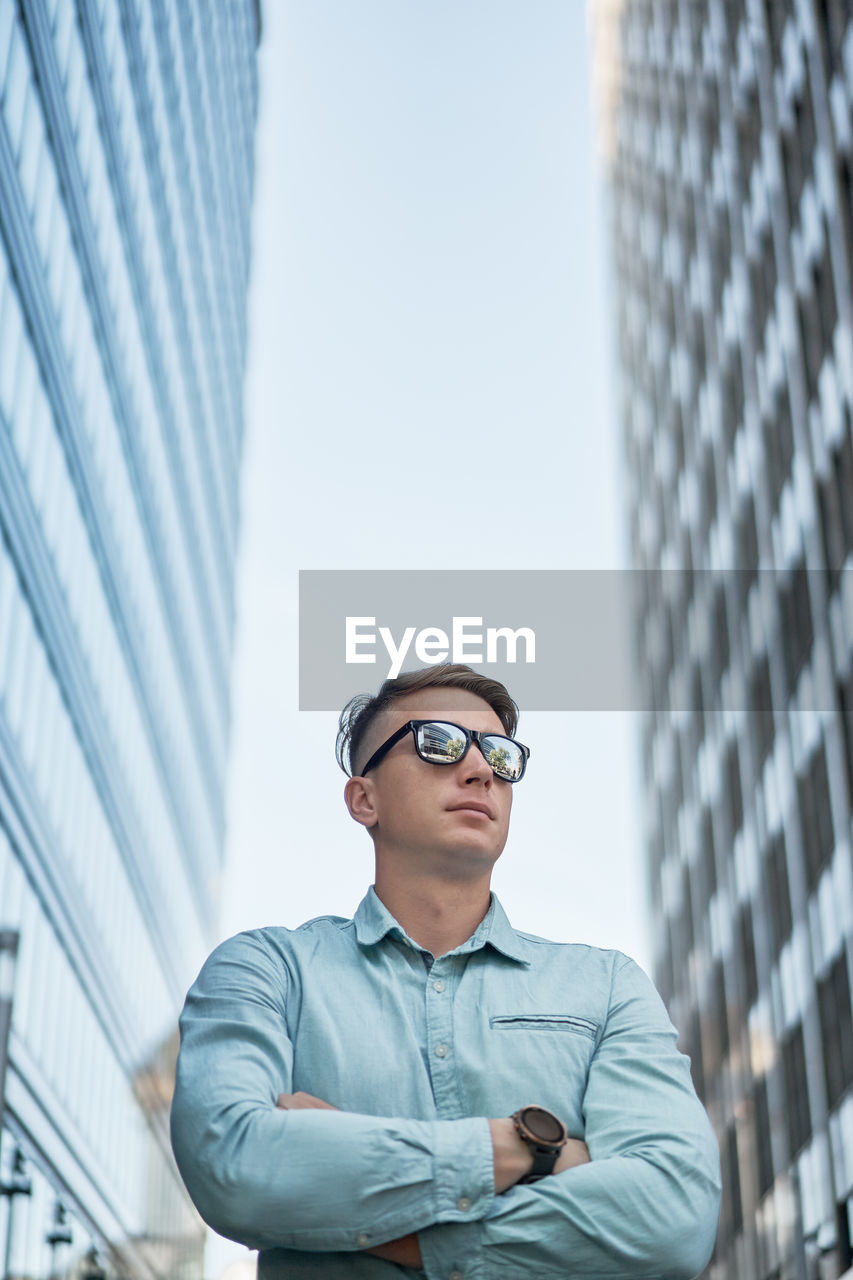 Low angle of serious businessman with sunglasses standing in downtown looking away while waiting for meeting in city