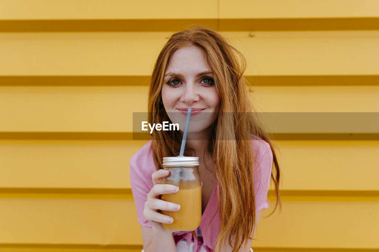 Smiling woman holding smoothie in front of yellow wall