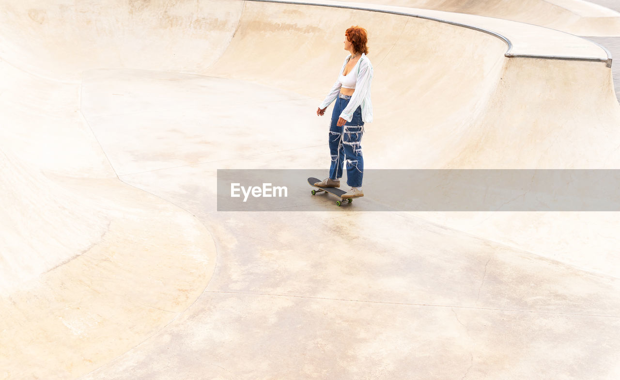 From above full body side view of active female in casual wear riding skateboard in skate park with ramp on summer day