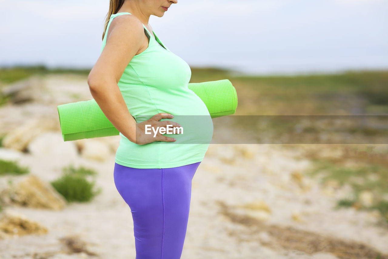 Midsection of pregnant woman with exercise mat standing on land