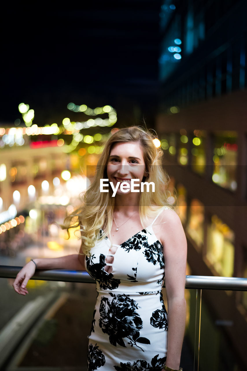 Portrait of smiling young woman standing in balcony at illuminated city