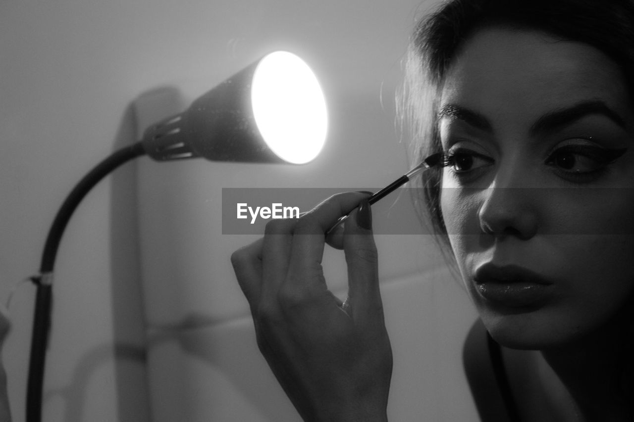 Close-up of young woman applying mascara by illuminated lamp