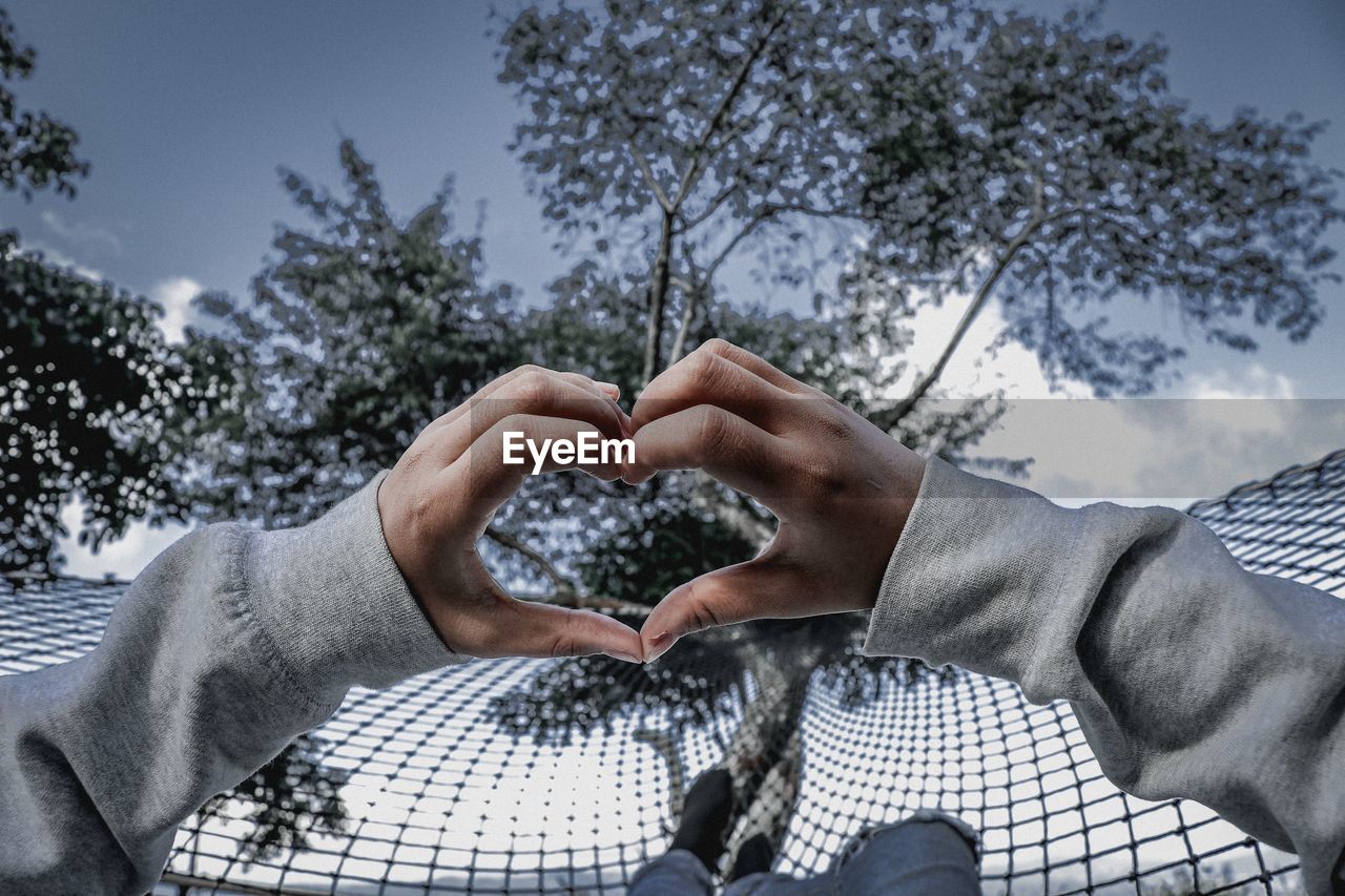 MIDSECTION OF MAN MAKING HEART SHAPE AGAINST TREES AND SKY