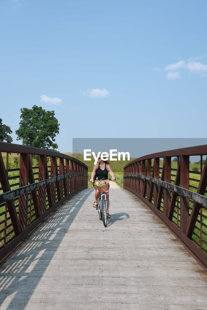 Full length of mature woman riding bicycle on footbridge against blue sky during sunny day