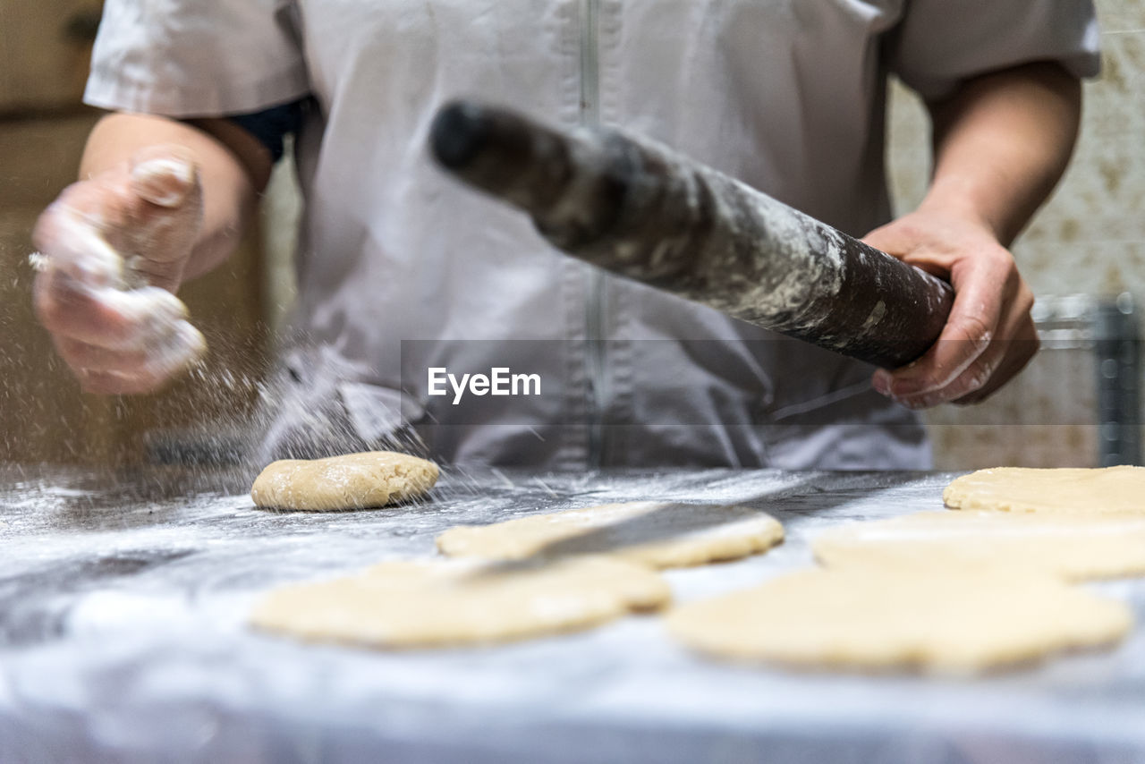 Unrecognizable baker with rolling pin spilling flour on table while preparing pastry in kitchen