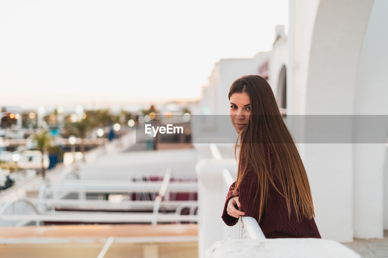 Portrait of young woman standing in balcony against clear sky