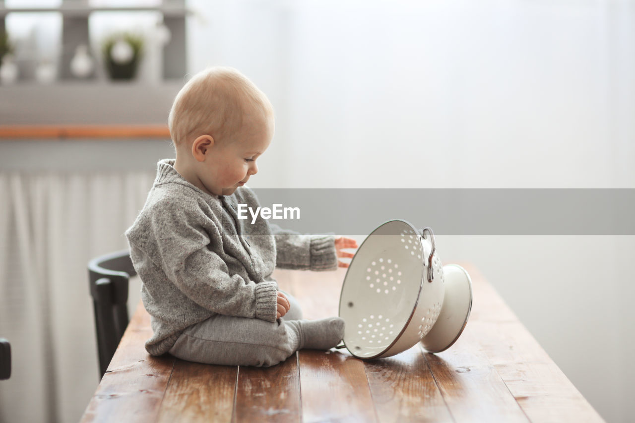 Baby of 10 months plays on the table in the kitchen with kitchen utensils