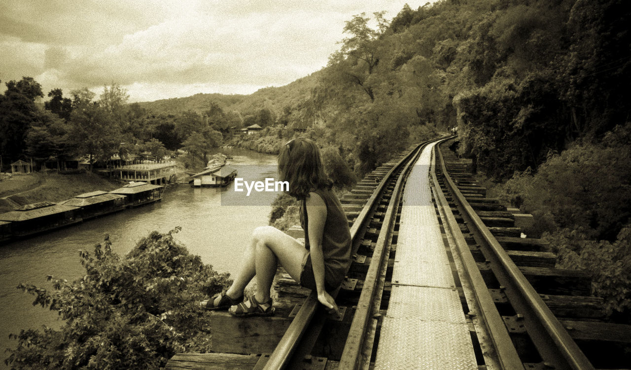 Full length of woman sitting on railroad track by river against sky