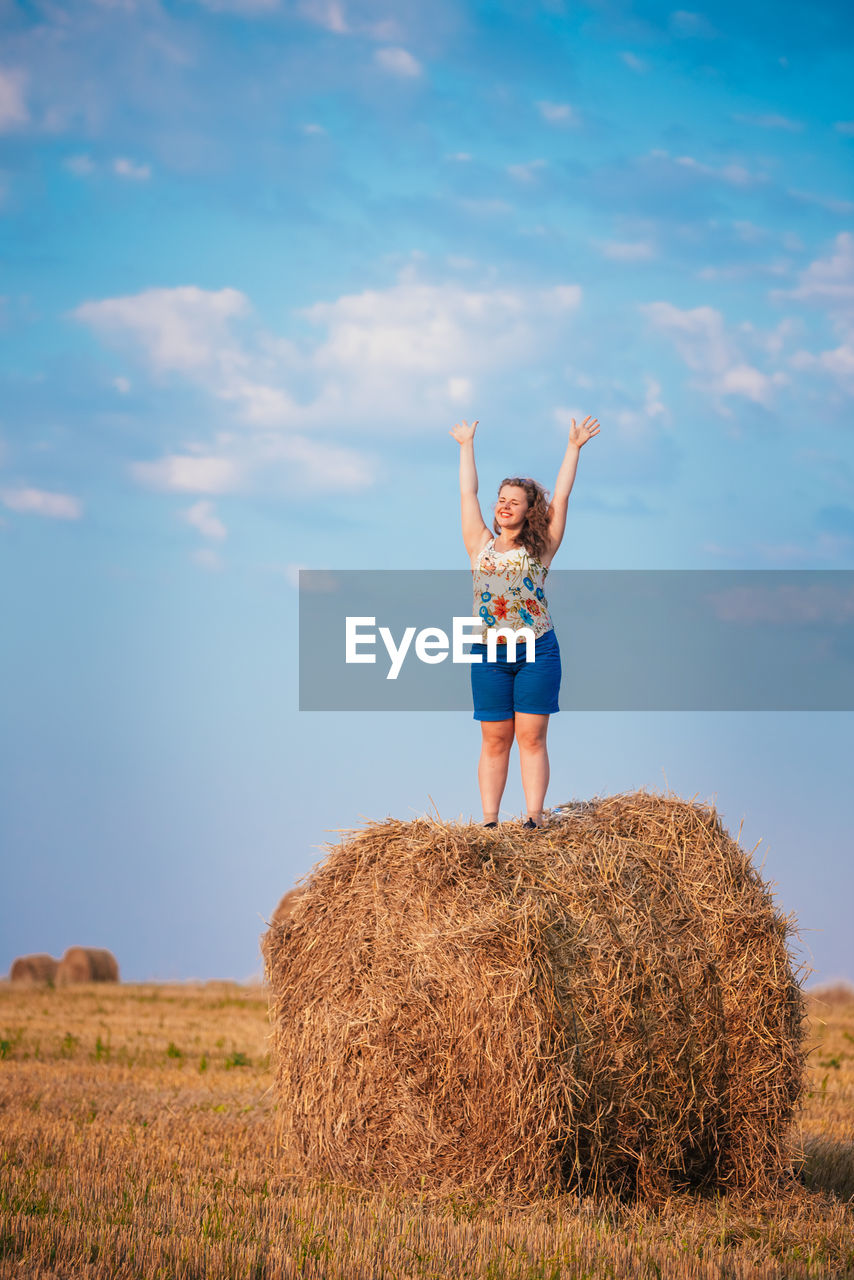 Woman with arms raised standing on hay bale against sky