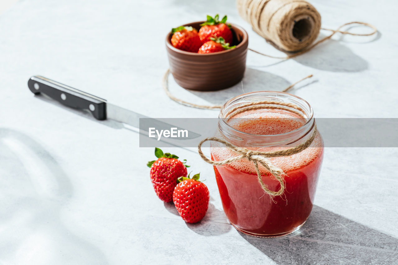 From above composition with fresh homemade strawberry juice in glass jar wrapped with twine placed on marble surface with whole berries and knife