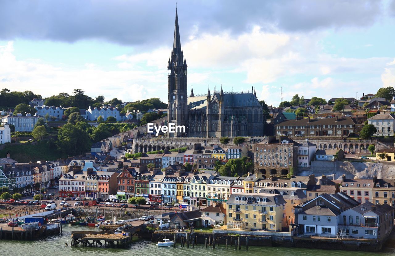 St colmans cathedral amidst buildings by river against cloudy sky during sunny day