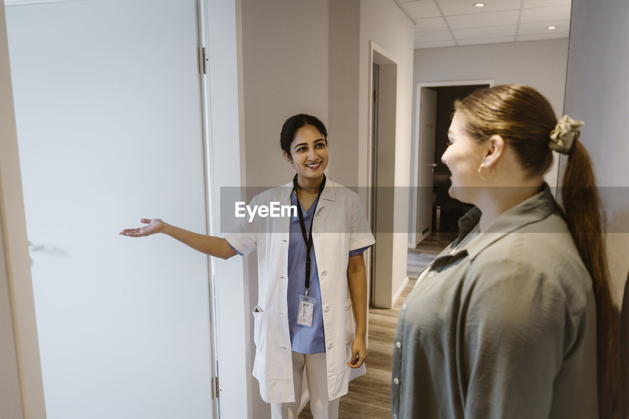 Smiling doctor assisting female patient towards medical examination room at clinic