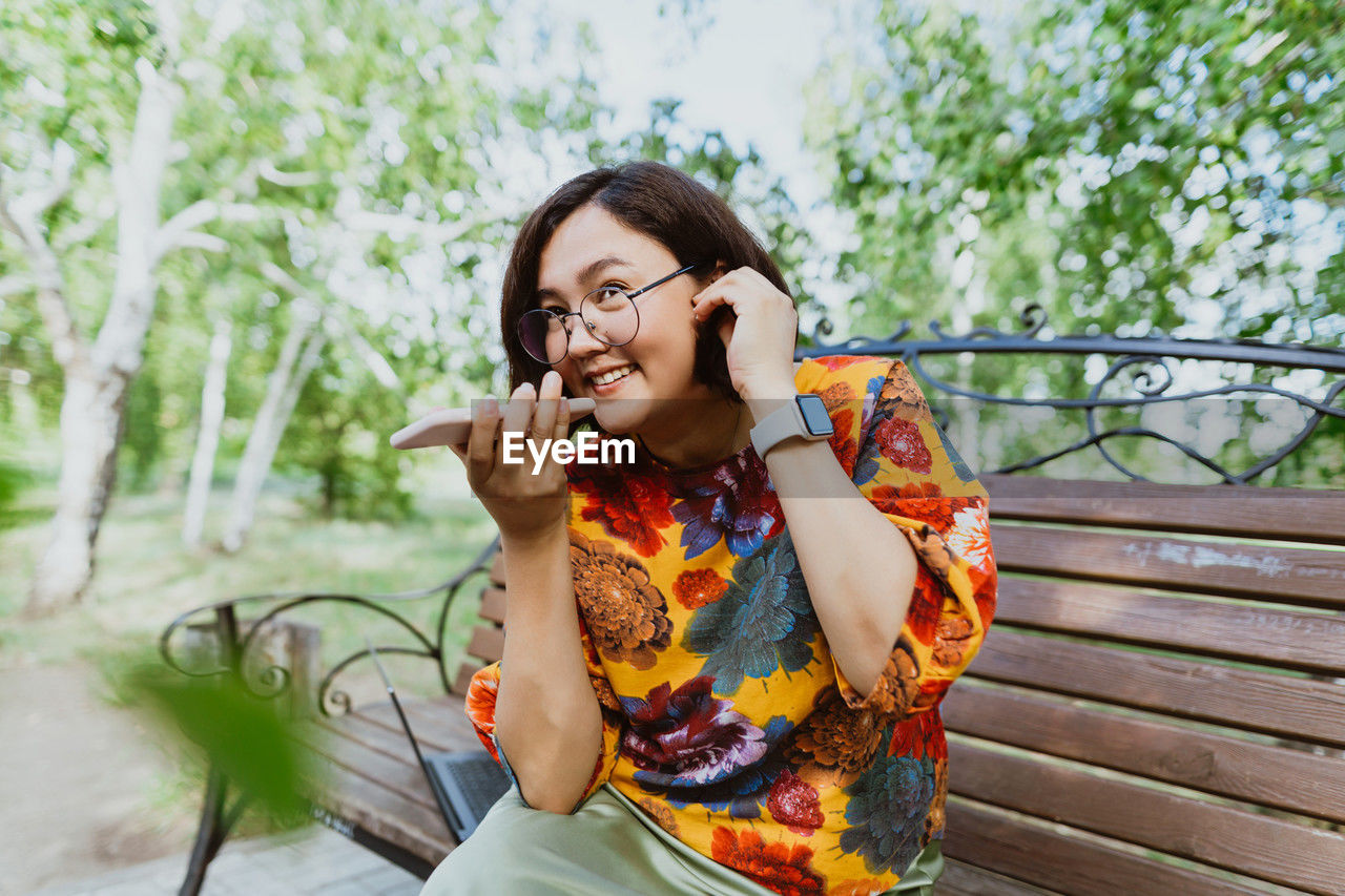 Happy woman sitting on bench in a green park, emotionally talking on her phone