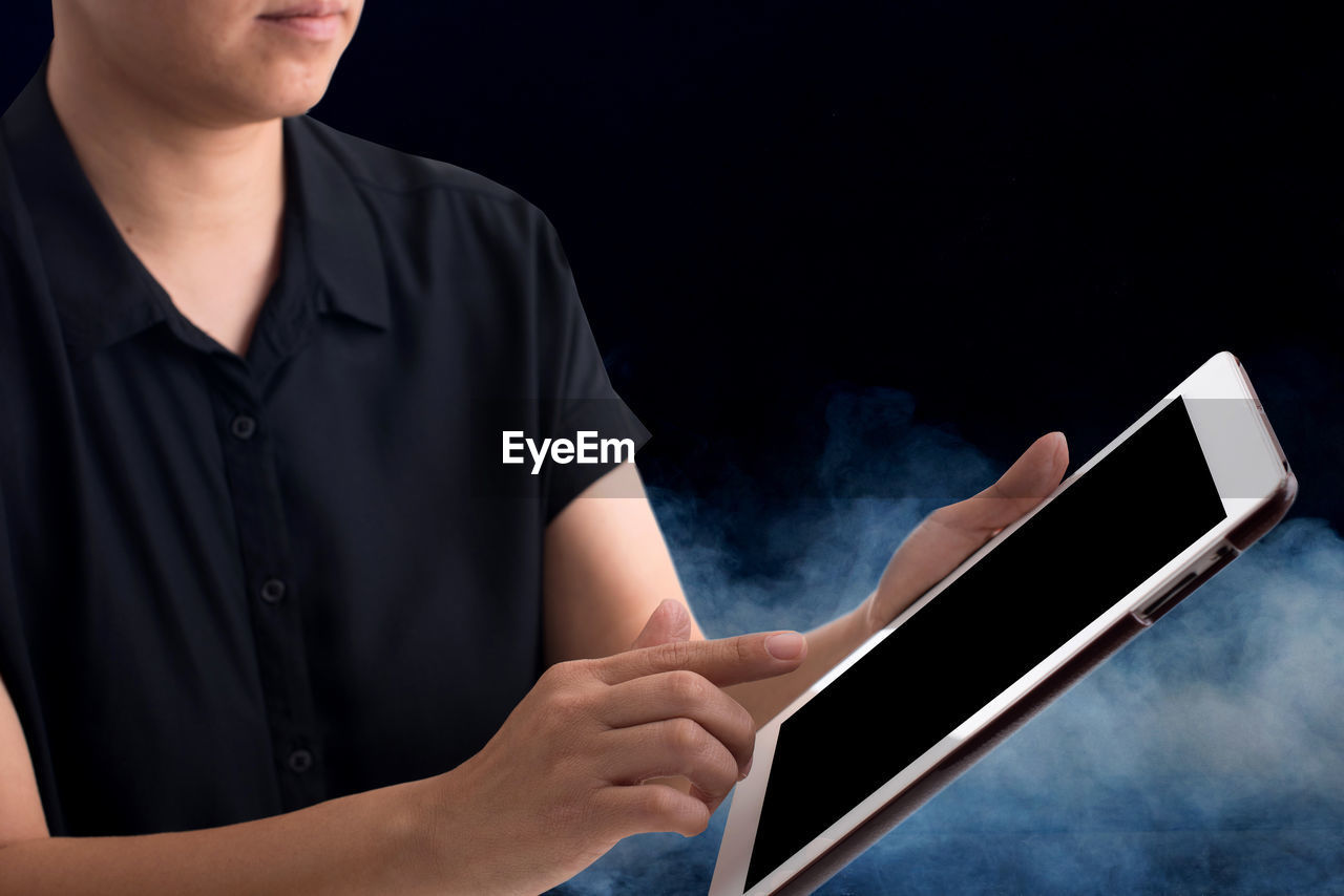 Midsection of woman using digital tablet standing amidst smoke against black background