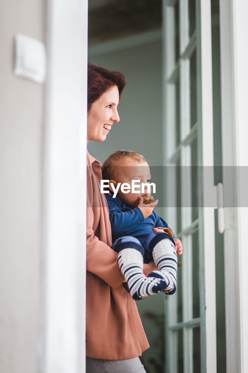 Smiling mother with son looking away while standing at home entrance