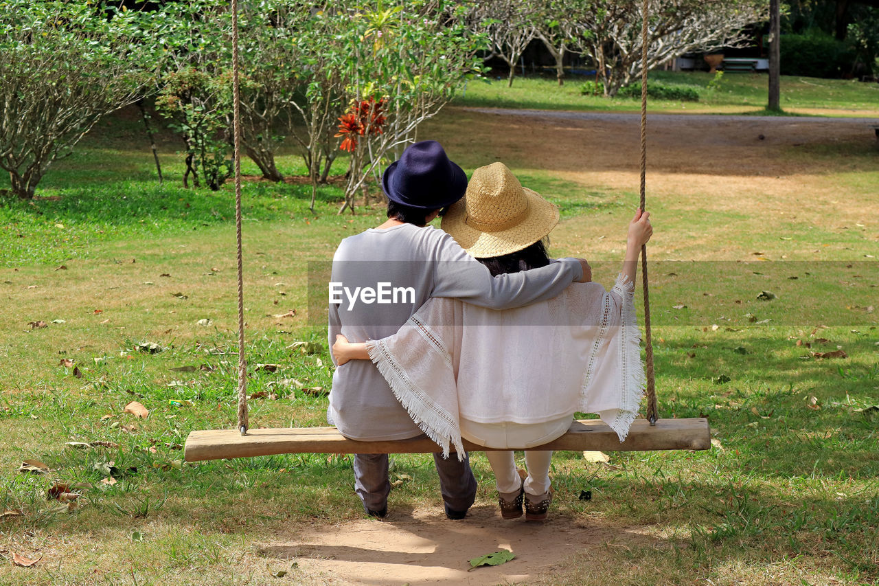 Rear view of couple in hats sitting on swing in park
