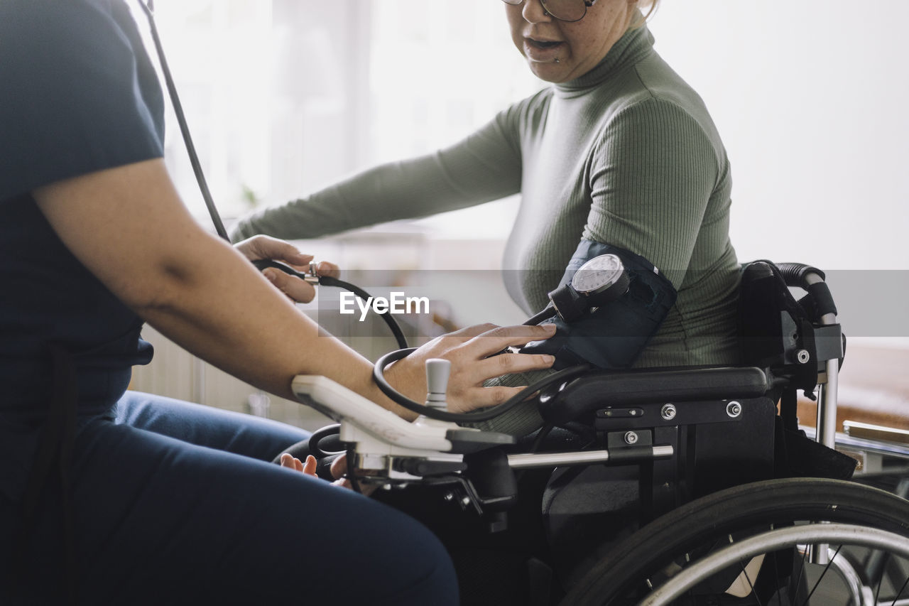 Midsection of female nurse checking blood pressure of woman sitting on wheelchair in clinic