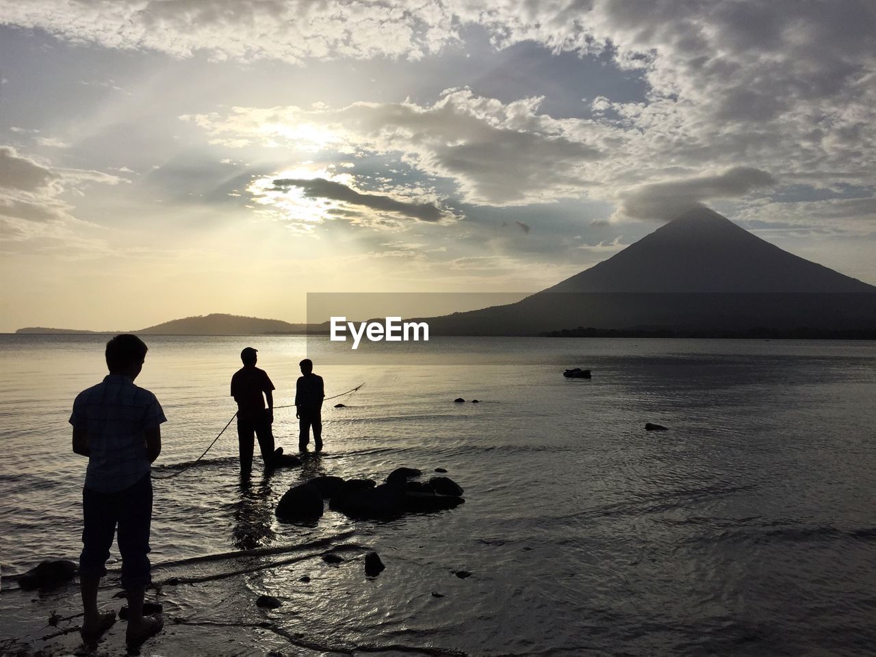 Silhouette men fishing in lake in front of mountains against cloudy sky during sunset