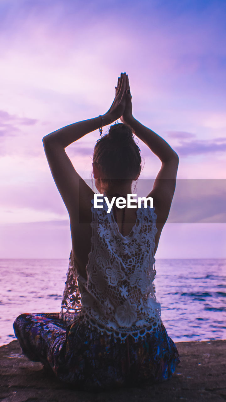 Rear view of woman with arms raised meditating at beach against sky during sunset