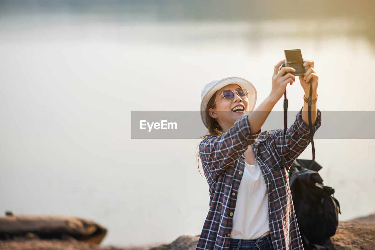 A female tourist happily takes a selfie by a reservoir with a camera.