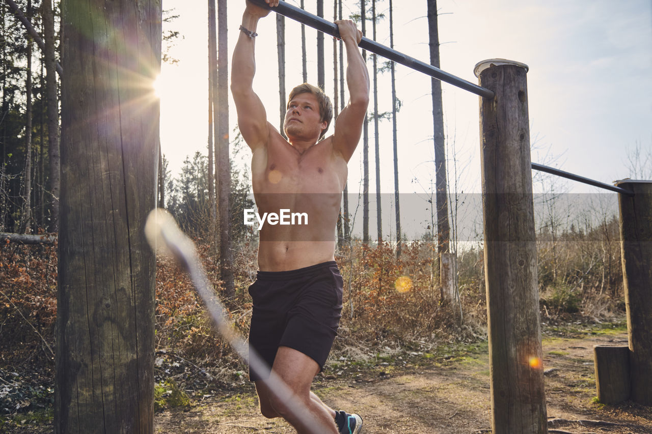 Shirtless male athlete hanging on horizontal bar on fitness trail in forest