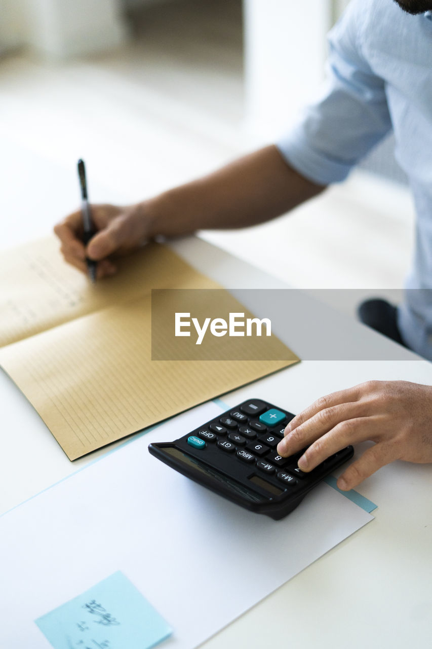 Businessman writing in book while using calculator at office