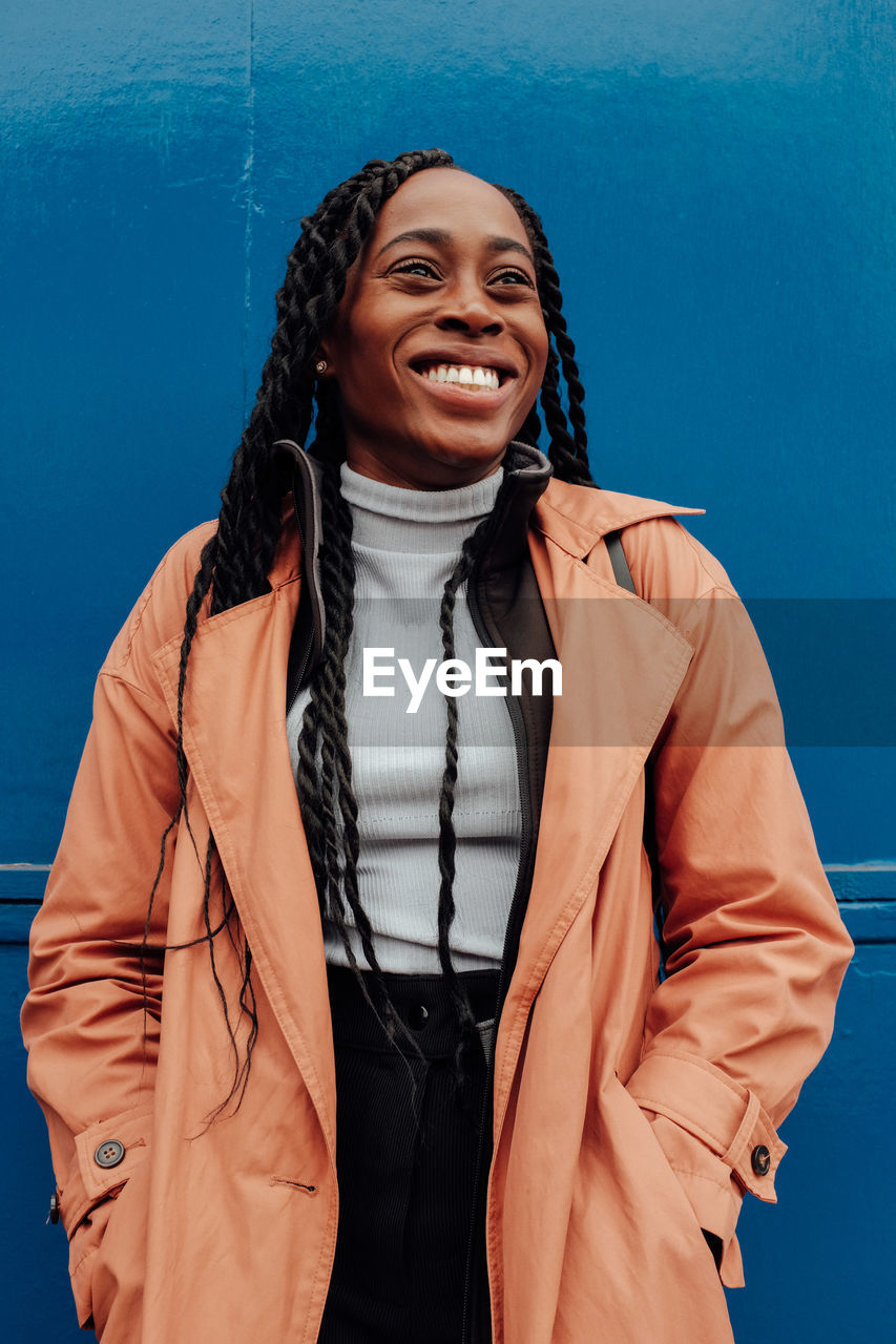 Smiling young woman wearing overcoat while standing against blue wall