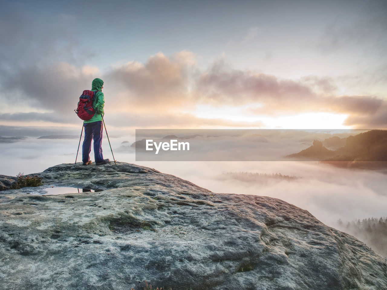 Tourist in green windcheater with hood on head and red backpack on back enjoy wild misty viee