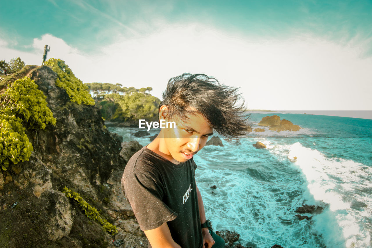 Portrait of young man standing by cliff against sea