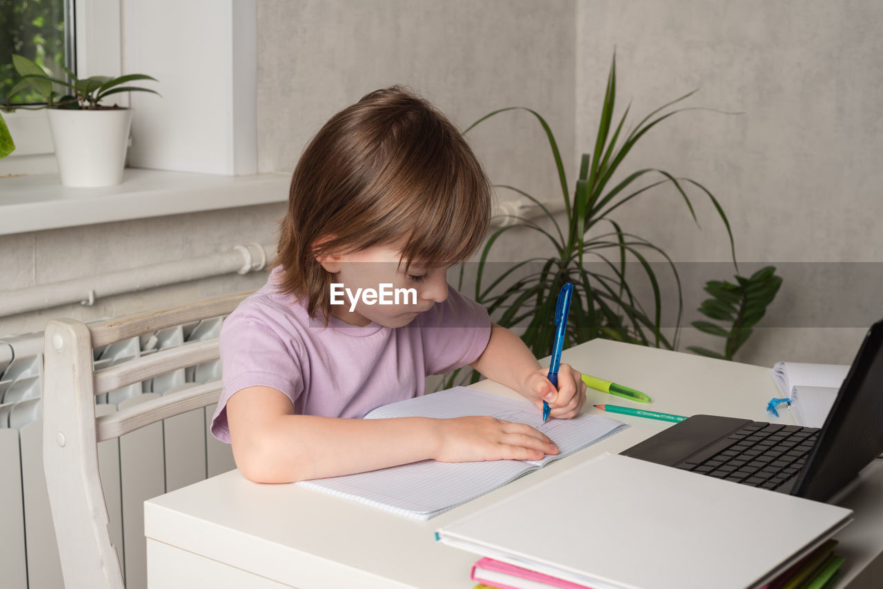 Happy girl sits at a table and works on a laptop and writes with her left hand. left handed child. 