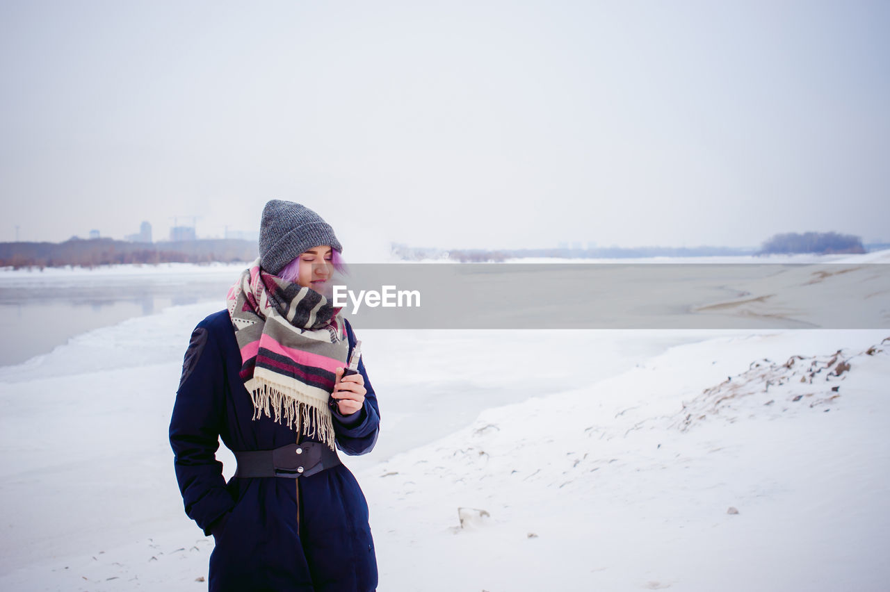 Woman smoking electronic cigarette while standing on snow covered landscape against sky