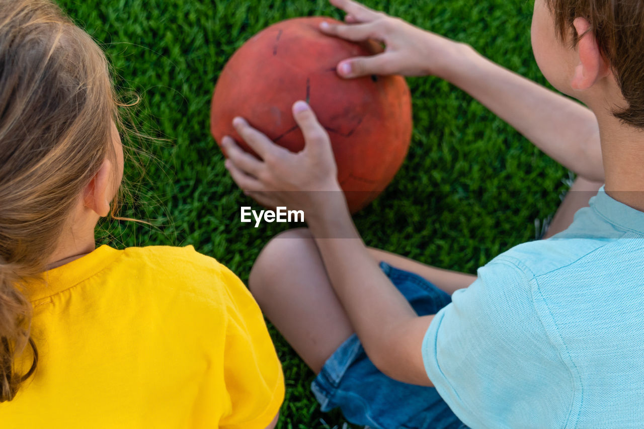 Unrecognizable children sitting on green grass and holding ball outdoors