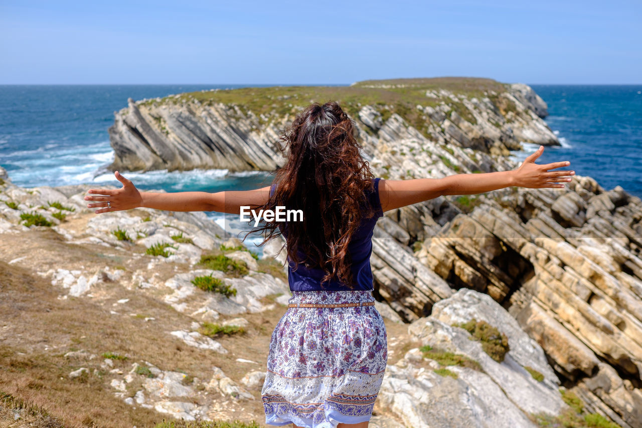 Rear view of woman standing on coast with her arms outstretched