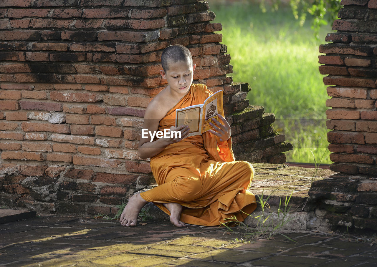 Monk reading book while sitting by brick wall