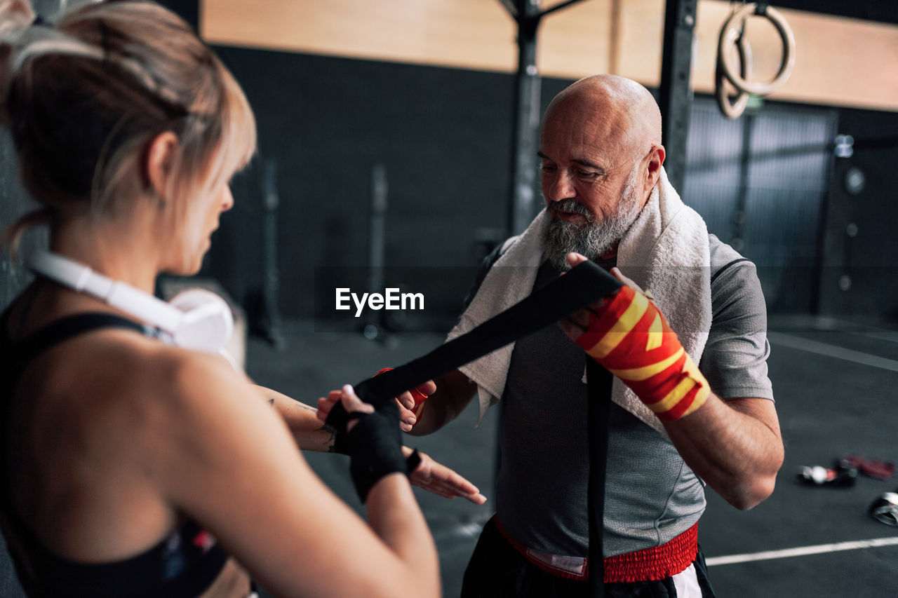Adult bearded male instructor helping woman athlete wrapping bandage on wrist while standing preparing for exercising in boxing gym