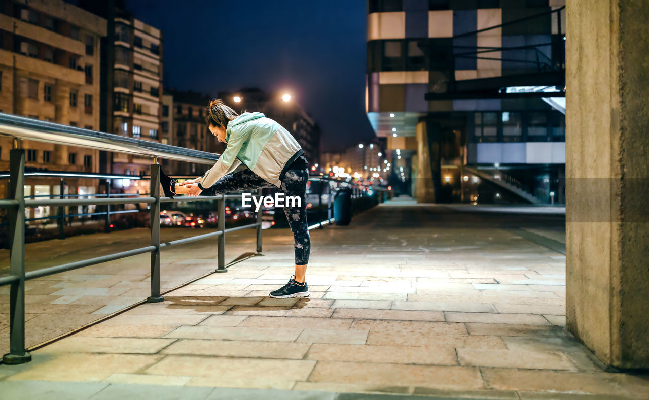 Woman runner stretching her legs over banister before training at night on town