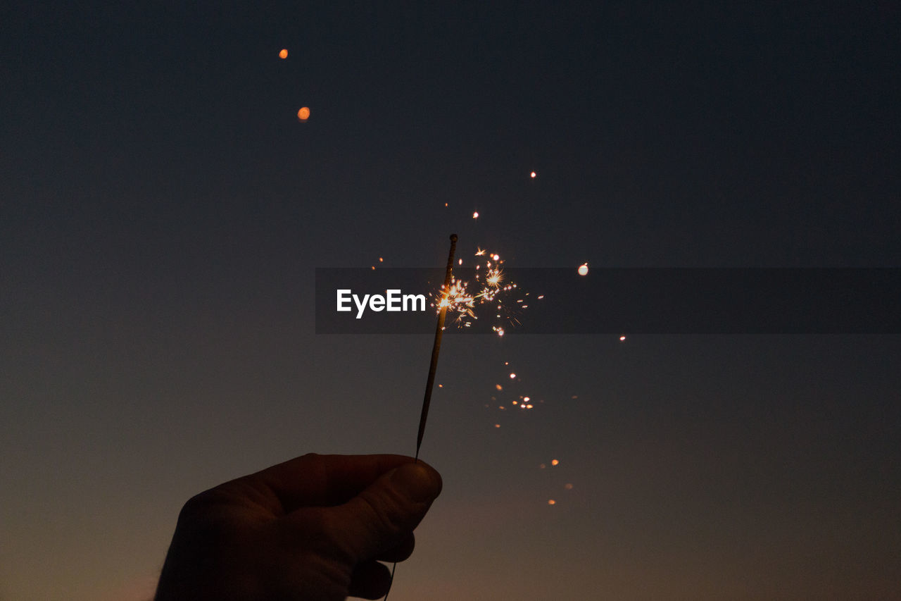 Cropped hand holding lit sparkler against sky at dusk