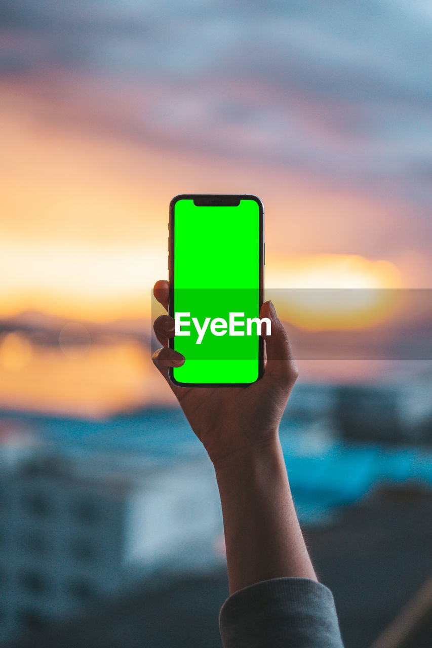 CLOSE-UP OF HAND HOLDING SMART PHONE AGAINST SUNSET SKY