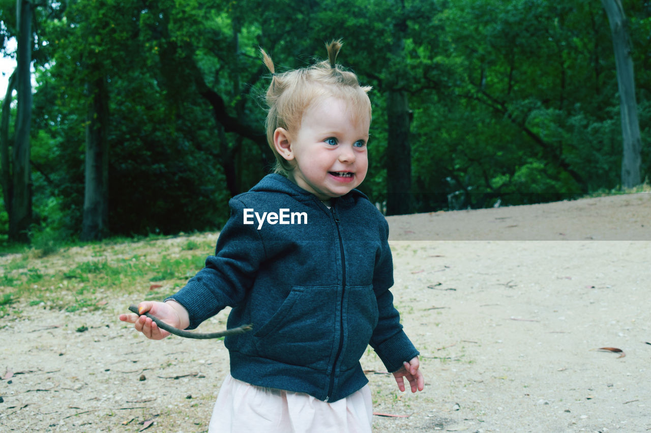 Baby girl holding stick while looking away against trees on field