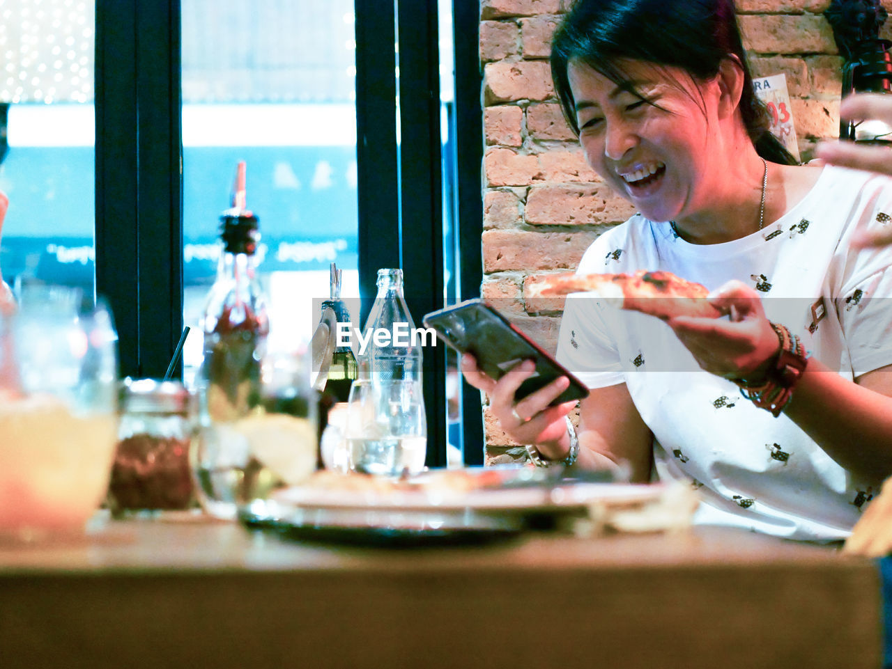 Cheerful woman using phone while having pizza at table in restaurant