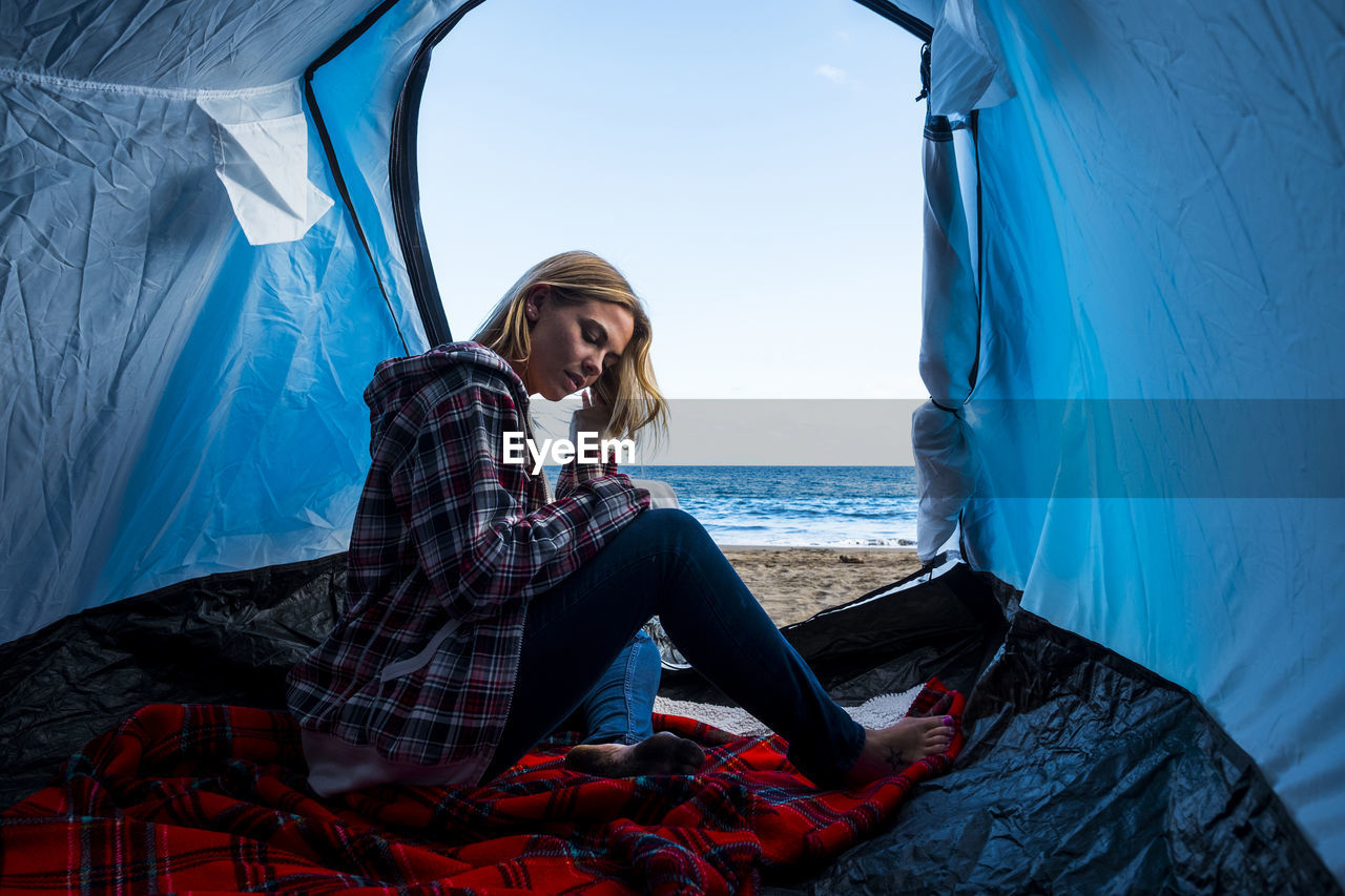 Woman sitting in tent at beach against sky