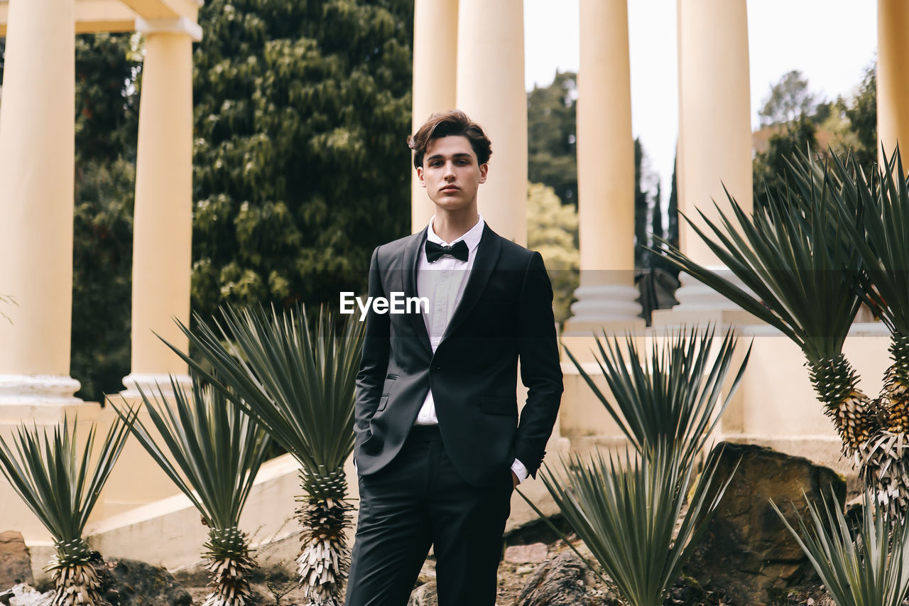 A beautiful young man, the groom in an elegant wedding suit, stands posing in the city's old park