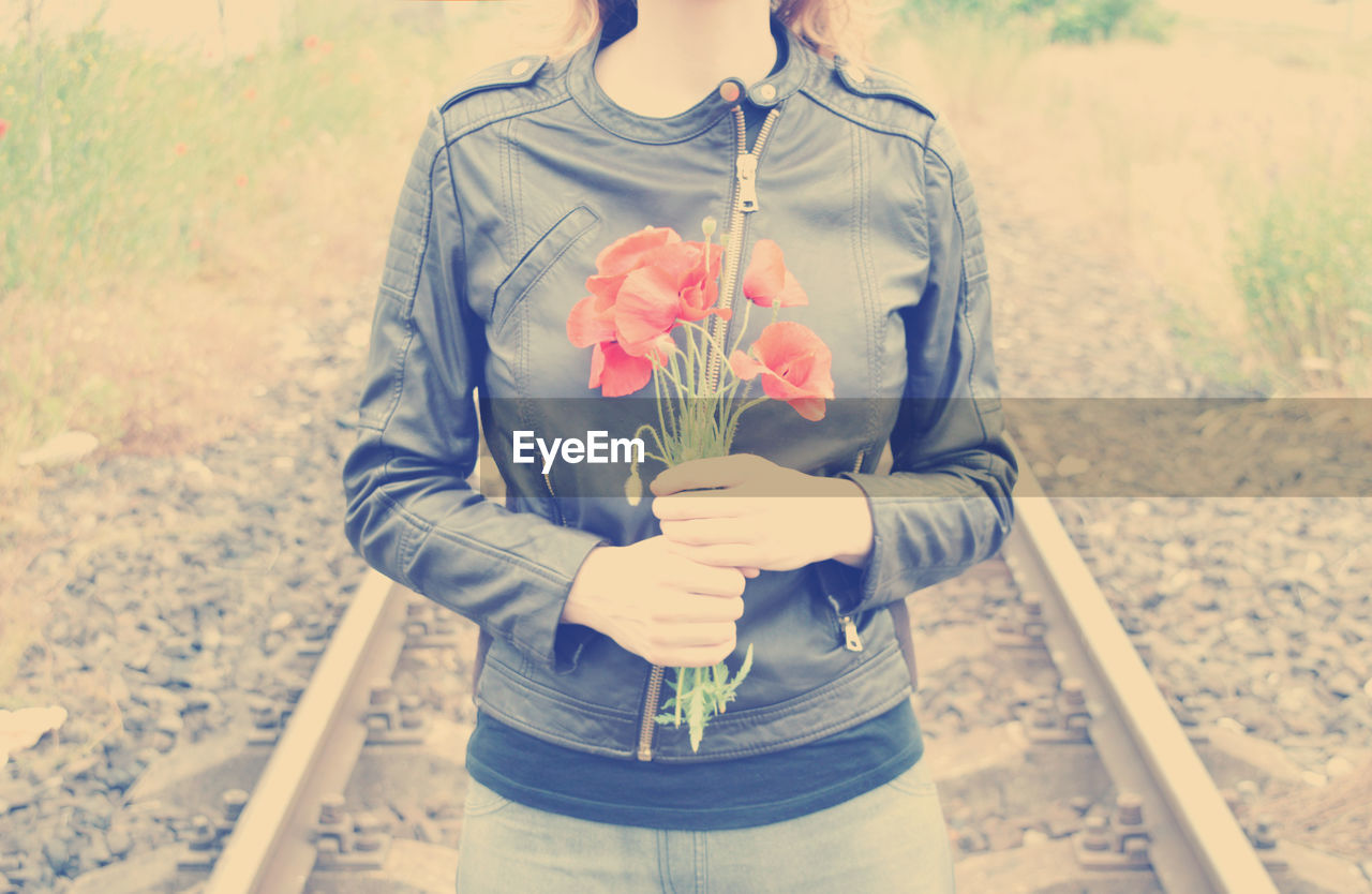 Midsection of woman holding red poppies while standing on railroad track