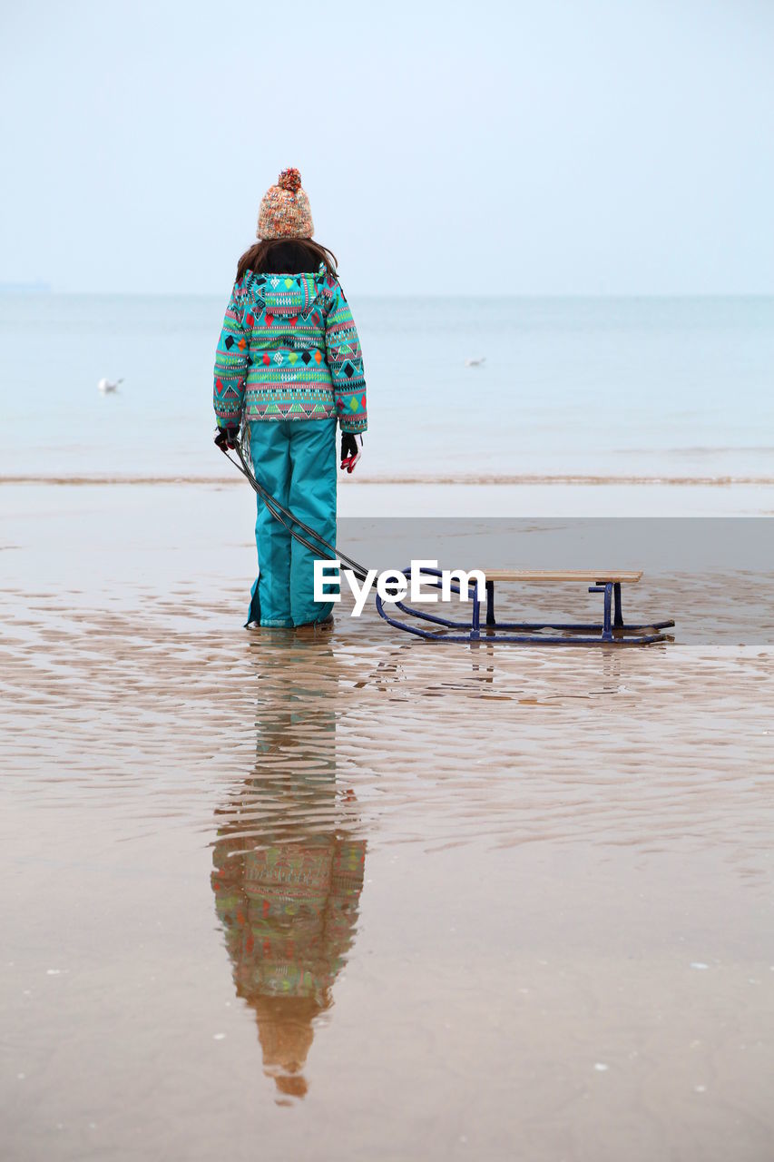 Reflection of girl with sledge standing at beach during winter