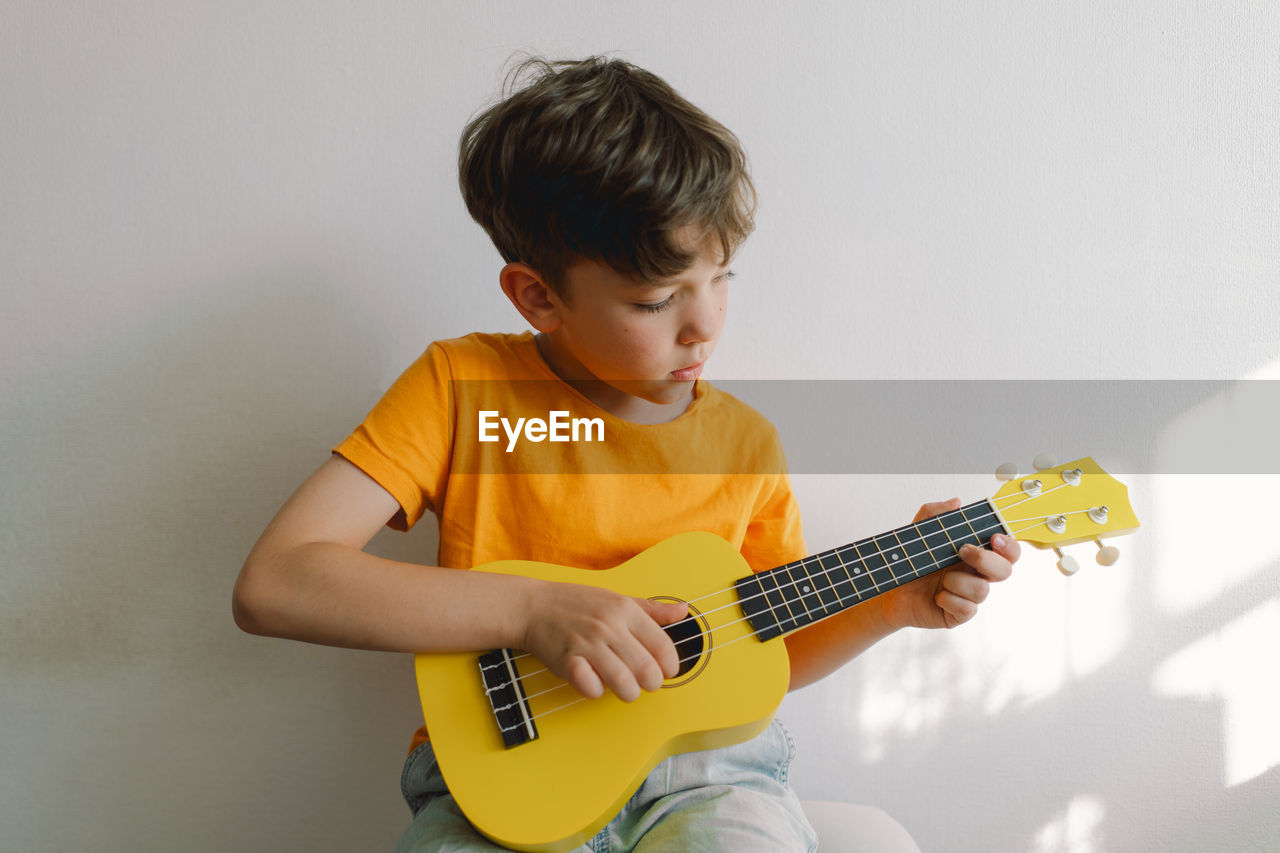 Cute boy learns to play the yellow ukulele guitar in home. cozy home.